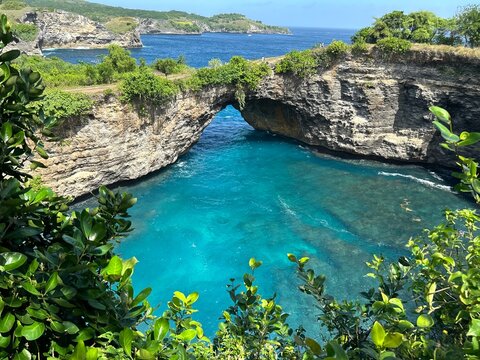 Scenic view of a natural rock arch over a turquoise ocean cove surrounded by lush green cliffs. - Powered by Adobe