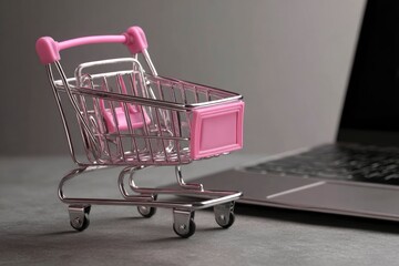 A mini pink shopping cart sits beside a laptop on a gray surface