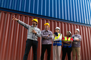 Logistics team leader pointing while diverse engineering staff observe and discuss in front of cargo containers. Concept of leadership, teamwork, and global trade operations