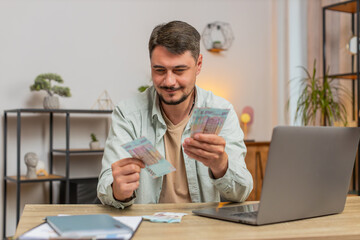 Planning budget. Rich happy man counting money Ukrainian hryvnias UAH cash using laptop calculate domestic bills at home table. Guy satisfied of income salary earnings money for planned vacation gifts
