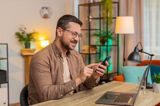 Happy young Caucasian man sitting at table, think and using mobile phone smiling at home apartment. Guy freelancer texting share messages on smartphone social media applications online watching movie.