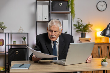 Senior freelancer accountant working with financial graph documents, calculate taxes for charts and reports at home office desk. Businessman grandpa working on laptop computer planning expenses budget