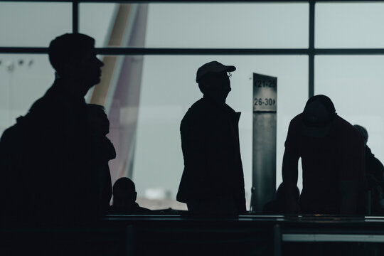 Silhouettes of diverse travelers waiting at an airport terminal with large windows and flight information displays