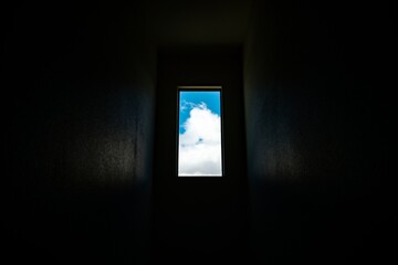 View of bright blue sky with white clouds through a narrow rectangular window in a dark interior space creating a contrast of light and shadow