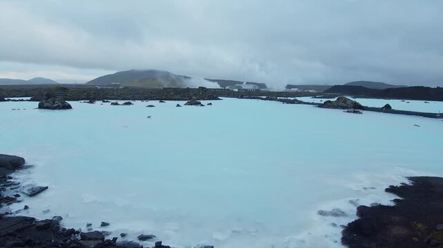 An aerial or wide shot of the famous Blue Lagoon geothermal spa in Iceland, showing the milky blue water surrounded by dark volcanic lava fields. In the background, the Svartsengi Geothermal Power Pla