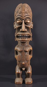 Tall carved wooden tribal statue with geometric patterns closed eyes open mouth and prominent phallus on dark background