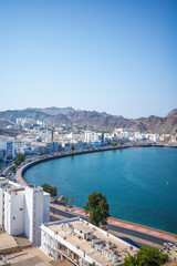High-resolution vertical photo of the Muttrah Corniche in Muscat, Oman, featuring a sweeping curved waterfront lined with white Arabian buildings, turquoise blue water, and rugged rocky mountains