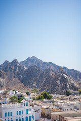 High-resolution vertical photo of Old Muscat, Oman, featuring rugged rocky mountains rising behind traditional white and beige Middle Eastern buildings. A historic watchtower