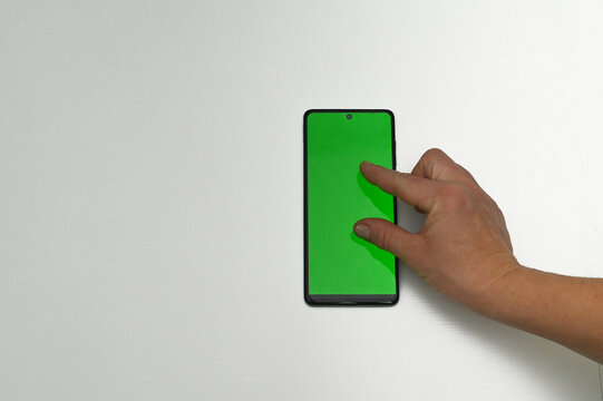 hand performs a gesture on the green smartphone screen, holding it vertically against a light background. smartphone with a green screen is used by a user who swipes or taps with his finger.