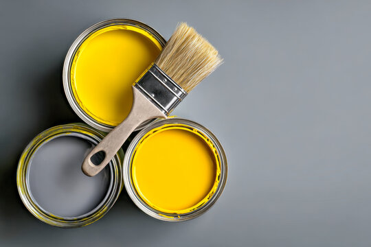 High angle view of three open paint cans with bright yellow and grey paint and a clean paintbrush resting on top against grey background