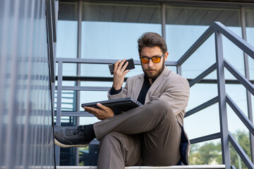 A man is sitting on the stairs while looking at his phone and holding a tablet