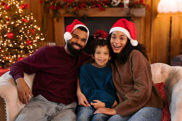 Happy African American family sitting on couch during Christmas time, wearing Santa hats and smiling at camera, surrounded by holiday decorations and twinkling tree