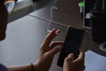 An Asian man wearing glasses is holding a cigarette and a cell phone in the office where he works in the morning.
