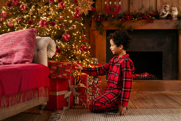 Happy black boy in red checkered pajamas kneeling by beautifully decorated Christmas tree, examining wrapped gifts. Happy New Year
