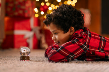 Happy Latin boy in red plaid shirt lying on the floor carpet, captivated by shimmering snow globe,...