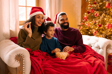 Excited Latin family sitting on couch wrapped in cozy blankets, enjoying holiday movie night,...