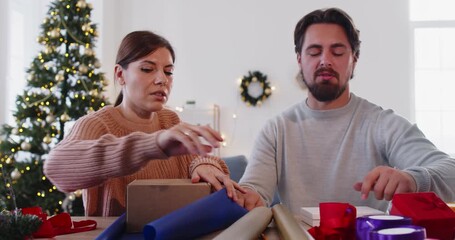 Christmas couple wrapping gift box at home with discussion. Talking man and woman measure paper, cut with scissors, and tie ribbon on presents in teamwork, sitting by Xmas tree and wreath on wall - Powered by Adobe