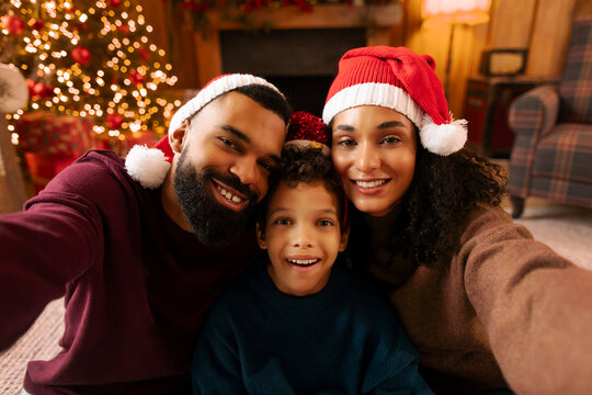 Happy Latin family taking selfie at Christmas Eve, wearing Santa hats, sitting in warm festive decorated living room