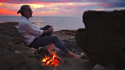 A contemplative man sits by a seaside fire at sunset, embodying mindfulness and reflection. The...
