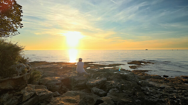 A solitary figure in white sits on rocky shores at sunset, facing the horizon. The scene evokes mindfulness and reflection, capturing a moment of solitude.