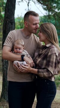 Happy young parents with their firstborn baby. Caucasian couple stand in nature with their newborn. Man kisses his wife on the forehead. Vertical video