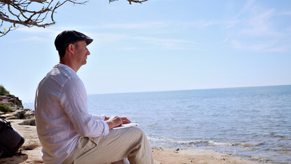 A focused digital nomad works on a portable device by the serene ocean, embodying freedom and remote work. The clear sky and tranquil waters highlight a peaceful, productive environment ideal. © stockcopter