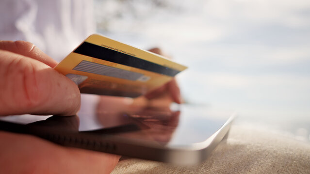 A digital nomad's hand holds a credit card for online payment, with a laptop nearby, symbolizing remote work's freedom and flexibility in a serene, possibly coastal setting. - Powered by Adobe
