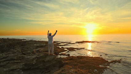 A person stands on rocky shores at sunset, arms raised in triumph. The golden sky and calm sea...