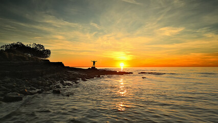 A serene coastal sunset scene with a lone statue on a rocky shore. The sun's golden glow over the calm sea evokes mindfulness and reflection, ideal for mental wellbeing and peace.