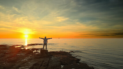 Person on rocky shore at sunset, arms outstretched, embracing the horizon. Serene ocean and vibrant sky evoke mindfulness and reflection, symbolizing peace and connection with nature.