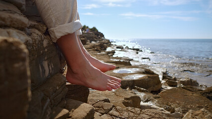 A person leans against a rocky cliff, bare feet dangling over the edge, overlooking the ocean. The serene coastal scene evokes mindfulness and reflection, capturing a moment of escape.