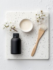 A minimalist flat lay composition featuring a black bottle, a small bowl of creamy liquid, a wooden brush, and delicate white flowers on a speckled tray.