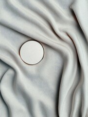 A close-up, top-down view of a round, white object with a thin gold border resting on softly draped, light gray fabric.