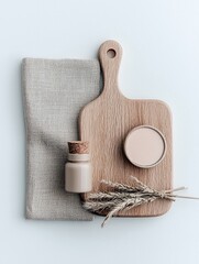 A minimalist arrangement of a wooden cutting board, beige fabric, a small bottle, a compact, and dried wheat stalks.
