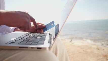 A digital nomad works on a laptop by the sea, holding a credit card, symbolizing remote work and financial transactions. The serene beach setting highlights the freedom and flexibility. - Powered by Adobe