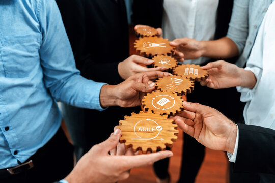 A diverse group of professionals engages in a collaborative effort, holding wooden gears that represent the agile process and effective workflow enhancement in business settings. Amity
