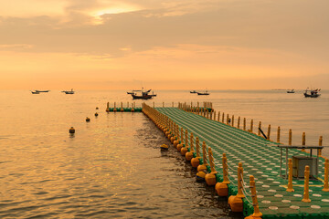 Floating dock extending into calm sea during golden sunrise with fishing boats on the horizon under a colorful sky