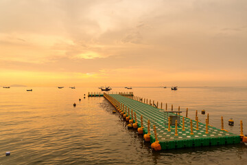 Floating dock extending into calm sea during golden sunrise with fishing boats on the horizon under a colorful sky
