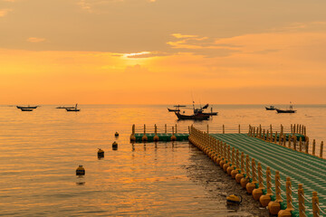 Floating dock extending into calm sea during golden sunrise with fishing boats on the horizon under a colorful sky