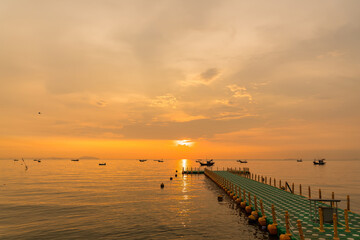Floating dock extending into calm sea during golden sunrise with fishing boats on the horizon under a colorful sky