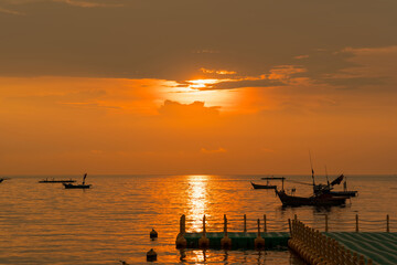 Fishing boats floating on calm sea during vibrant orange sunset with dock in foreground and golden reflections on water surface