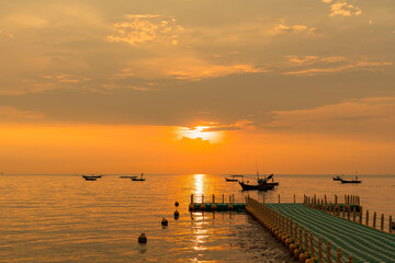 Floating dock extending into calm sea during golden sunrise with fishing boats on the horizon under a colorful sky