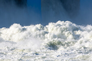 Huge white waves of rushing water splash and foam near the hydroelectric power plant dam gates during a massive flood discharge creating a dangerous chaotic natural abstract texture outdoors