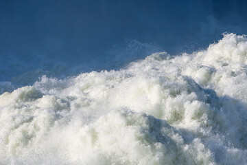 Strong powerful stream of rushing river water flows fast creating white foam and splashes against the deep blue background for a beautiful abstract natural texture of liquid energy outdoors