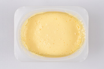 An overhead close-up shows a portion of light yellow, creamy cheese and vanilla dessert in a clear plastic container on a white background
