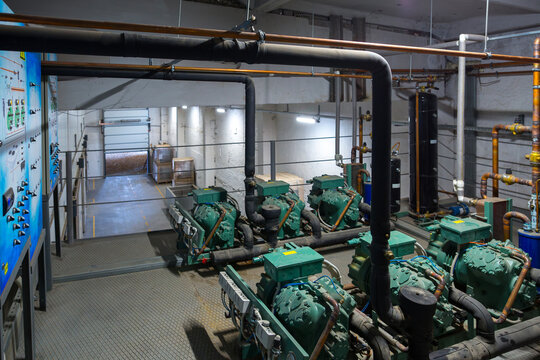 The industrial refrigeration plant room. Multiple green semi-hermetic compressors are connected to a system of copper and insulated pipes, tanks, gauges to provide cooling for a cold storage warehouse