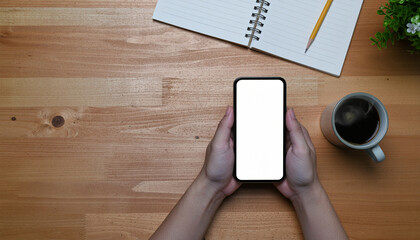 Top view of hands holding a smartphone with a blank white screen, perfect for a mockup, on a wooden desk with a notebook, pencil, and steaming coffee.