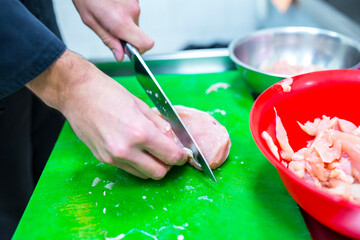 Close up view of a chef hands cutting raw chicken breast meat with a sharp knife on a green plastic board preparing fresh ingredients for pizza topping in the restaurant kitchen