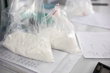 Three clear plastic bags filled with an unknown white powder or crystalline substance sit on a desk. The bags are on top of papers with handwritten notes possibly indicating illicit drugs or chemicals