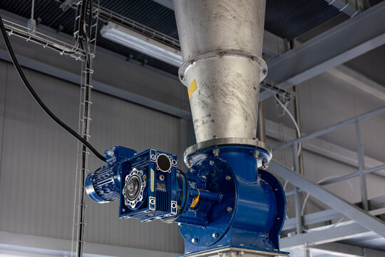 A blue industrial rotary valve, or airlock, is installed below a large stainless steel hopper. This equipment, with its electric motor and gearbox, is used for feeding bulk powder in a modern factory.